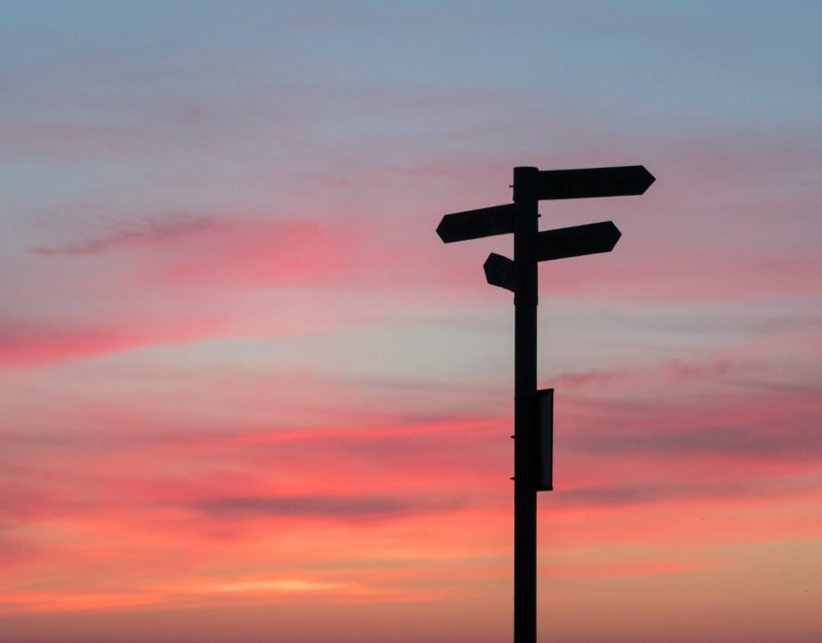 Wegweiser mit mehreren Richtungspfeilen vor einem Sonnenuntergang in Pastelltönen.