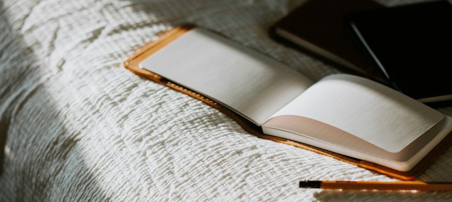 Offenes Notizbuch auf einer weißen Decke, daneben einige Stifte und ein Notebook.